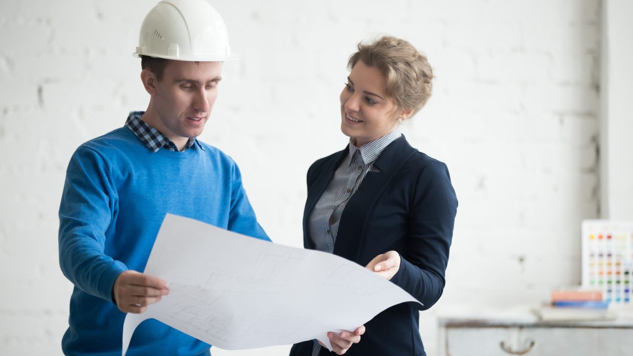 Architects with project in hands Two young colleagues at meeting looking at project concept, holding blueprints, planning work. Business discussion with construction engineer in hardhat in architectural agency office. Interior shot